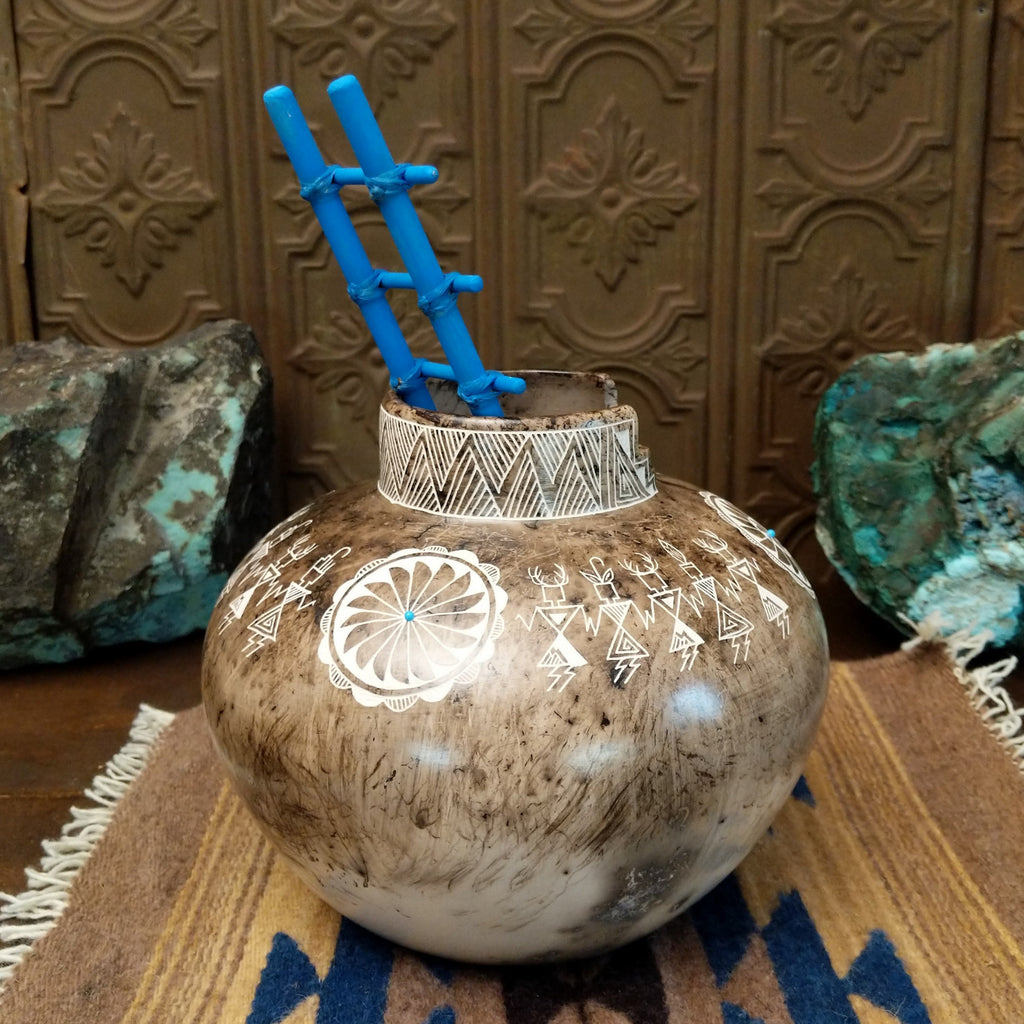 White clay pottery with fired horsehair and intricate etched designs of friendship dancers on the surface, stepped lip, and blue, wooden ladder.