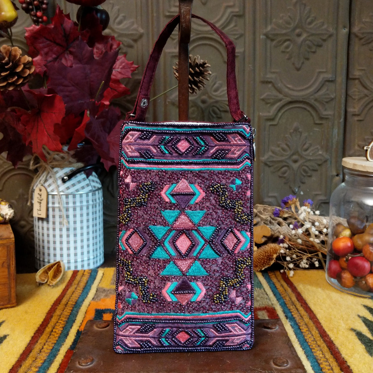 Aztec Club Bag by Bamboo Trading featuring intricate turquoise, pink, plum, and purple geometric patterns with diamond motifs, embroidered details, shimmering beadwork, sequins, and a coordinating burgundy shoulder strap.