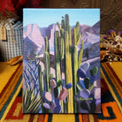 Bird Perch canvas artwork by Diana Madaras featuring towering green saguaro cacti, a small perched bird, colorful prickly pear plants, and purple desert mountains against a soft blue sky.