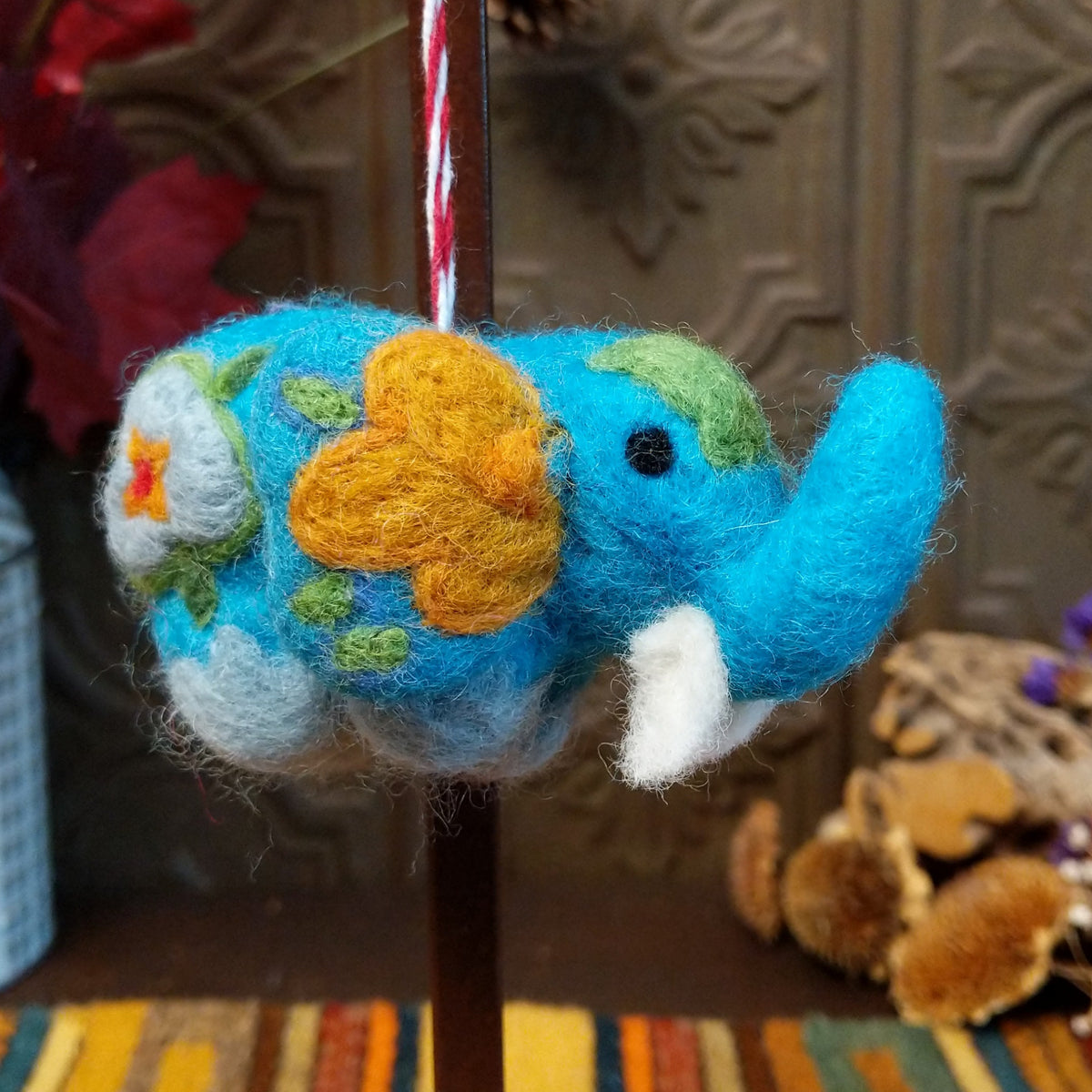 This colorful Blue Elephant Wool Ornament features a turquoise blue body, raised trunk, golden yellow flower, green leaves, white floral accents, black eyes, a white tusk, and a red and white twisted hanging cord.