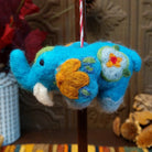This colorful Blue Elephant Wool Ornament features a turquoise blue body, raised trunk, golden yellow flower, green leaves, white floral accents, black eyes, a white tusk, and a red and white twisted hanging cord.