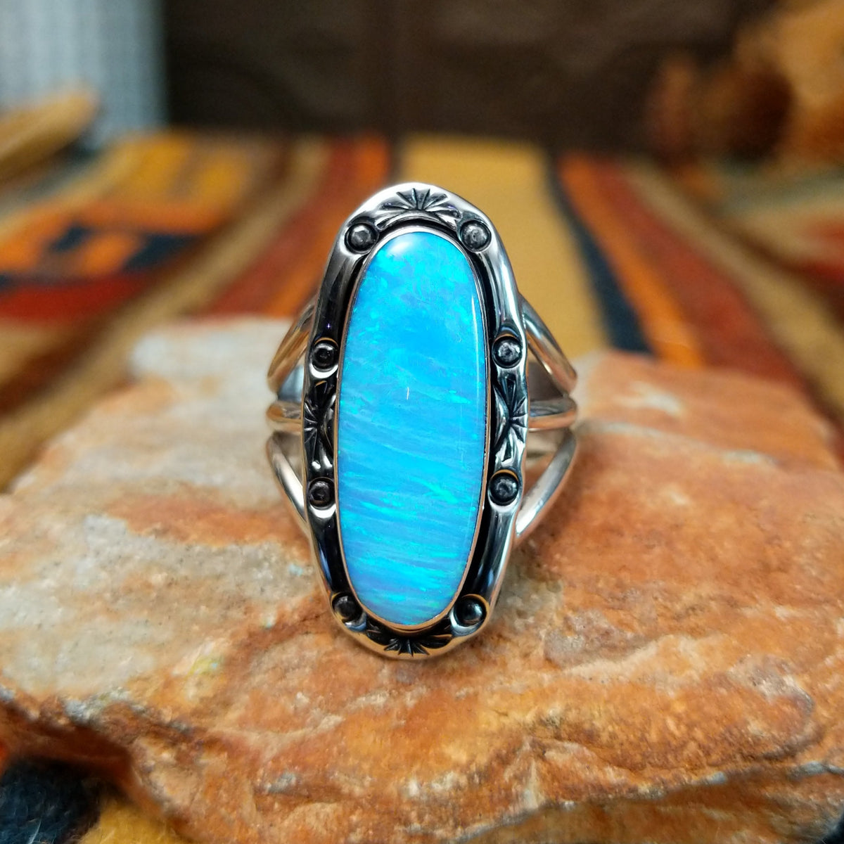 Blue Manmade Opal Ring by Terry Wood featuring an elongated oval blue opal with iridescent tones, decorative stamped sterling silver bezel, raised silver beads, and split-shank band.