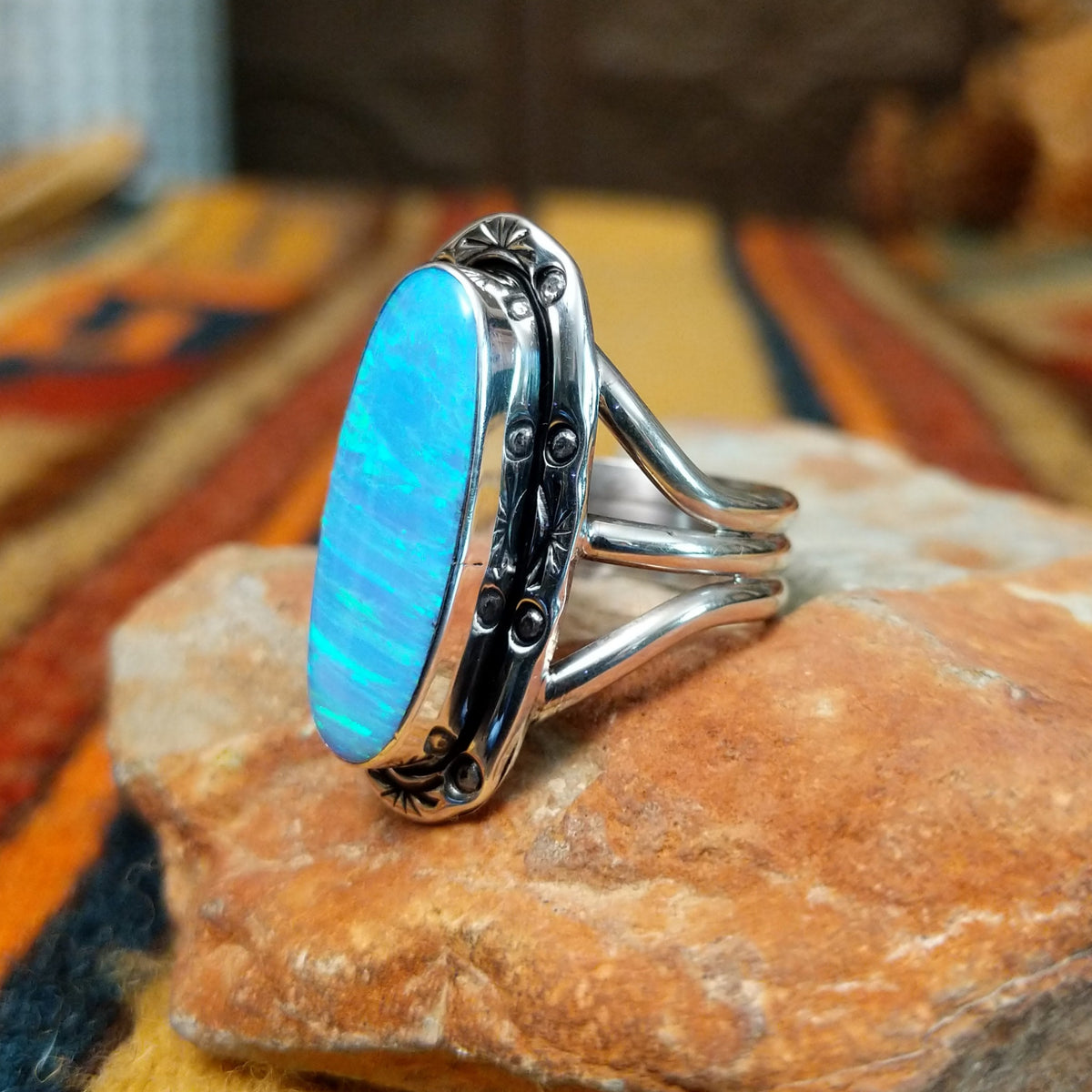 Blue Manmade Opal Ring by Terry Wood featuring an elongated oval blue opal with iridescent tones, decorative stamped sterling silver bezel, raised silver beads, and split-shank band; side view.