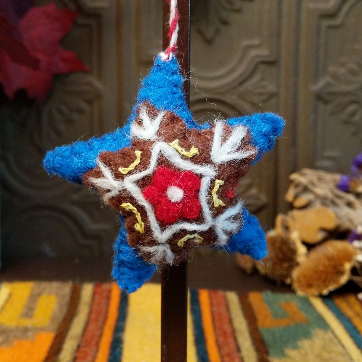 This charming Blue Star Wool Ornament features a vibrant blue five-pointed star with a brown center, white embroidered branches, yellow stitching, a red flower with a white center, and a red and white twisted hanging cord.
