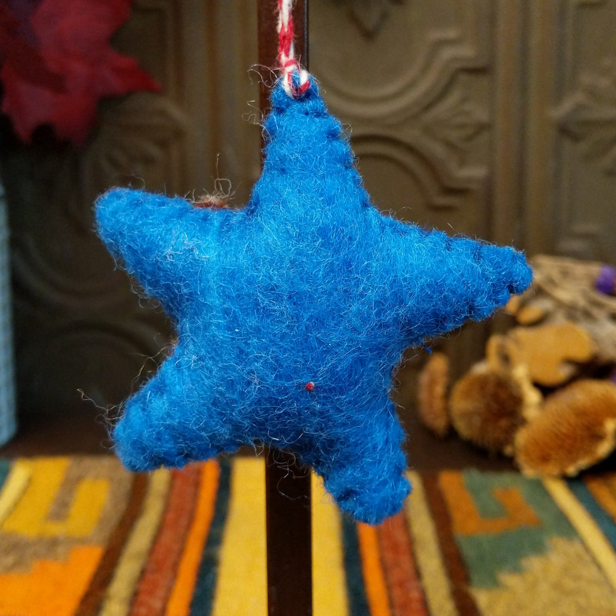 This charming Blue Star Wool Ornament features a vibrant blue five-pointed star with a brown center, white embroidered branches, yellow stitching, a red flower with a white center, and a red and white twisted hanging cord.