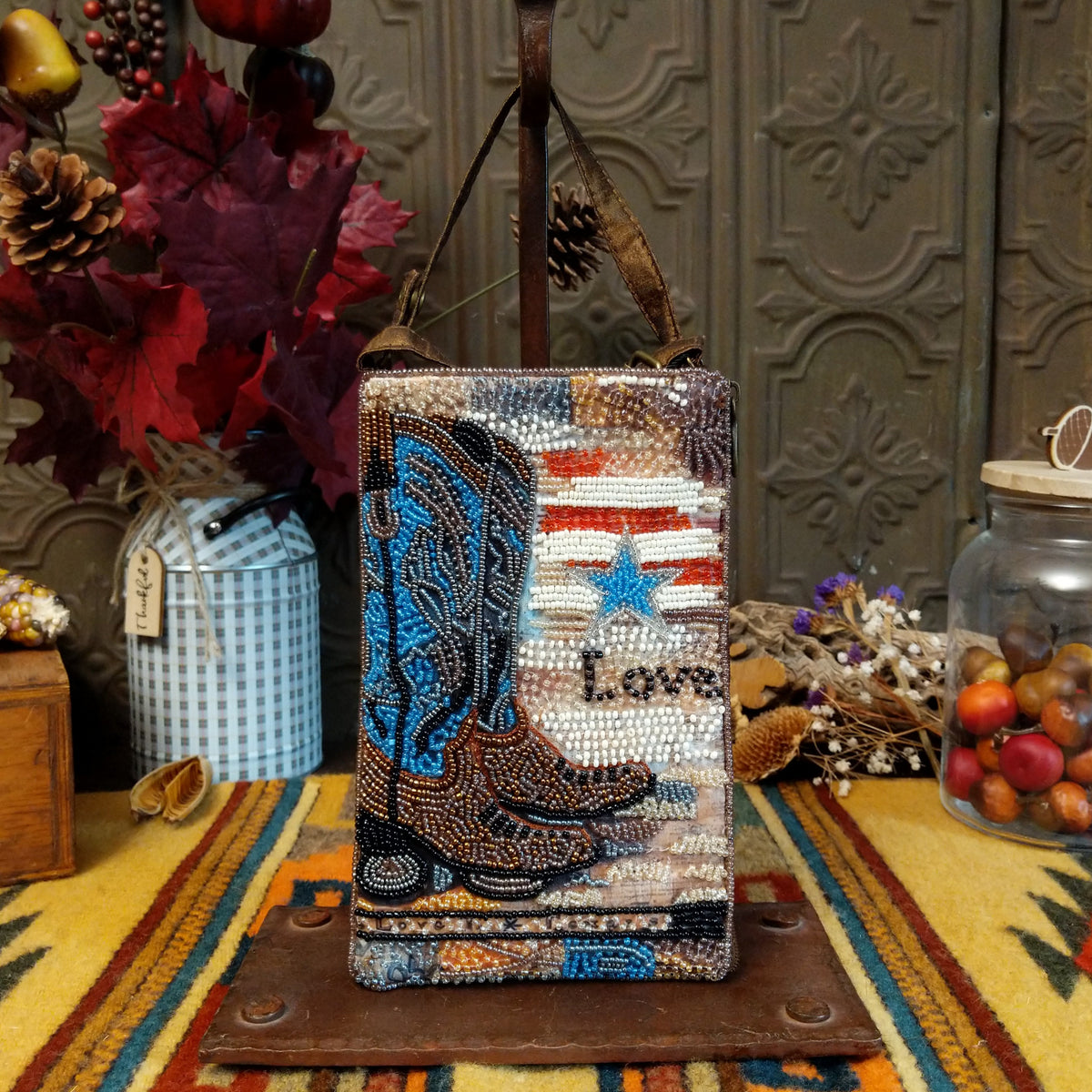 Boots & Spurs Club Bag by Bamboo Trading featuring intricately beaded brown and turquoise cowboy boots, a rustic American flag-inspired background, a sparkling blue star, and a brown shoulder strap.