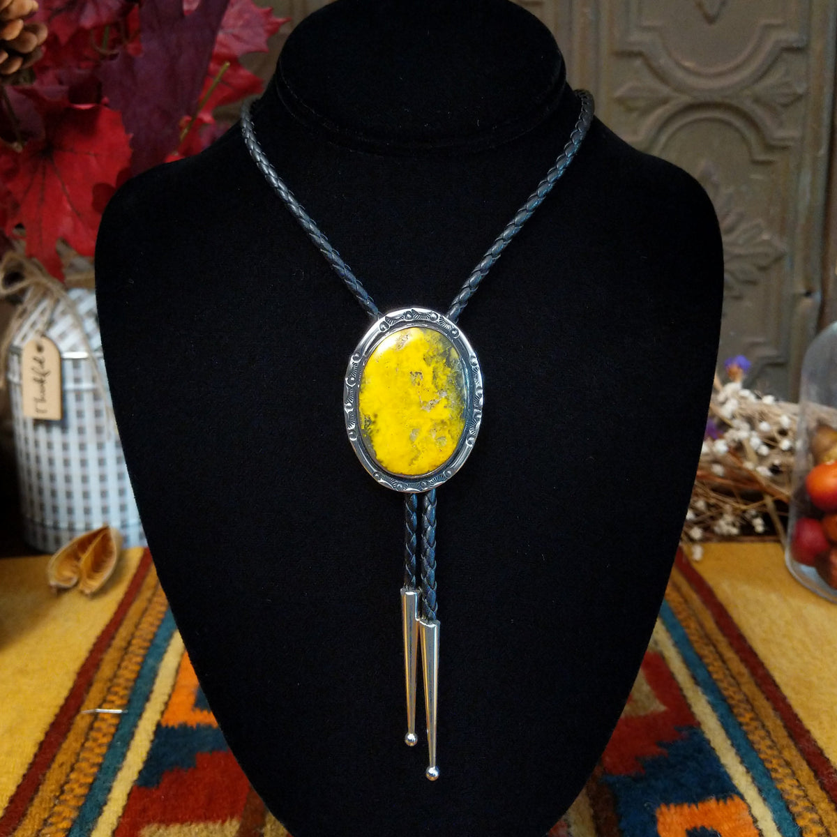 This striking Bumblebee Jasper Bolo Tie by Terry Wood features a bright yellow oval stone with gray and black matrix, a decorative stamped silver setting, braided black leather cord, and polished silver tips.