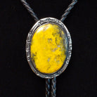 This striking Bumblebee Jasper Bolo Tie by Terry Wood features a bright yellow oval stone with gray and black matrix, a decorative stamped silver setting, braided black leather cord, and polished silver tips.