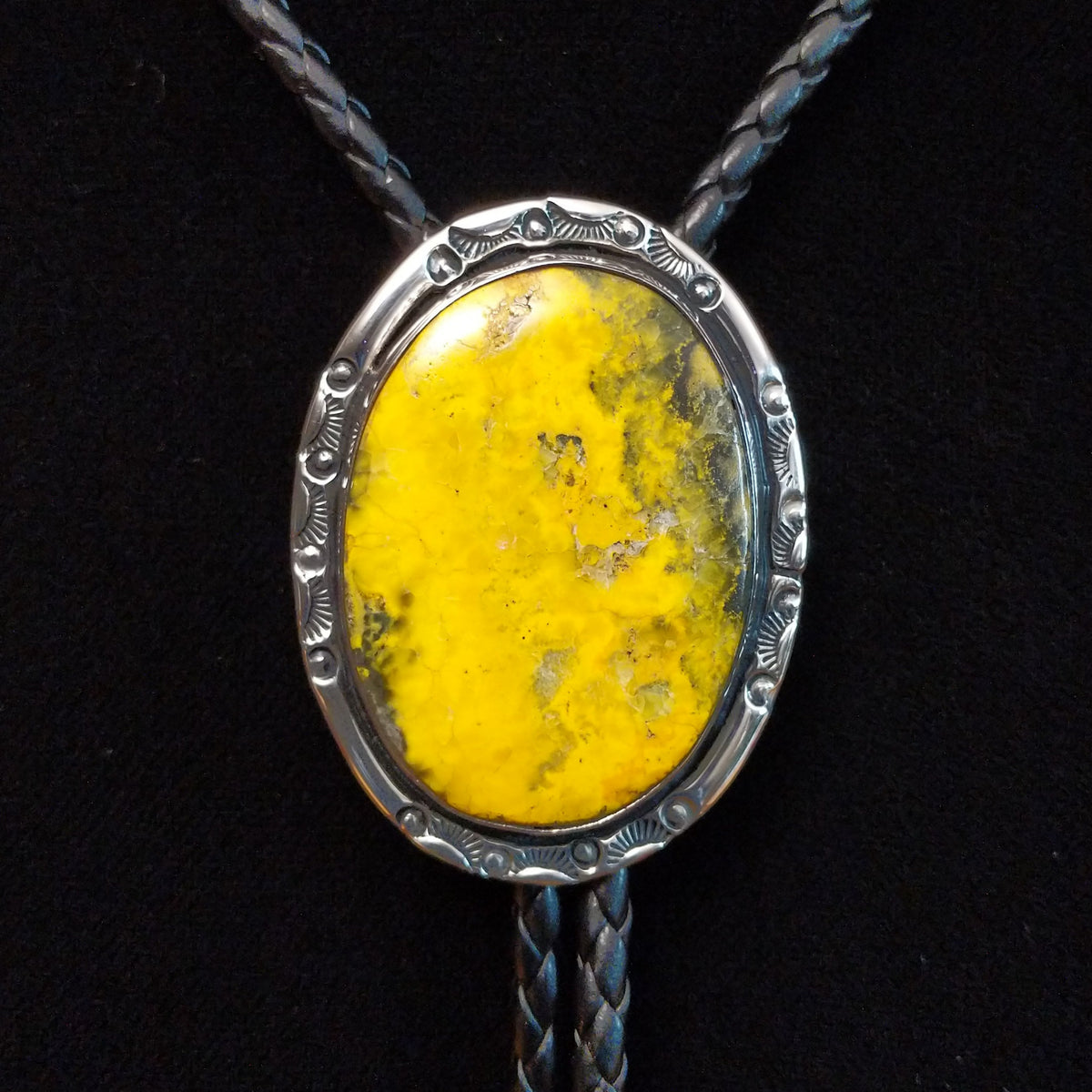This striking Bumblebee Jasper Bolo Tie by Terry Wood features a bright yellow oval stone with gray and black matrix, a decorative stamped silver setting, braided black leather cord, and polished silver tips.