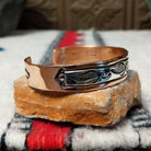 This Copper & Silver Feather Cuff by Robert Little features polished copper borders and a sterling silver center decorated with detailed feather motifs, curved accents, and dark oxidized stamping; side view.