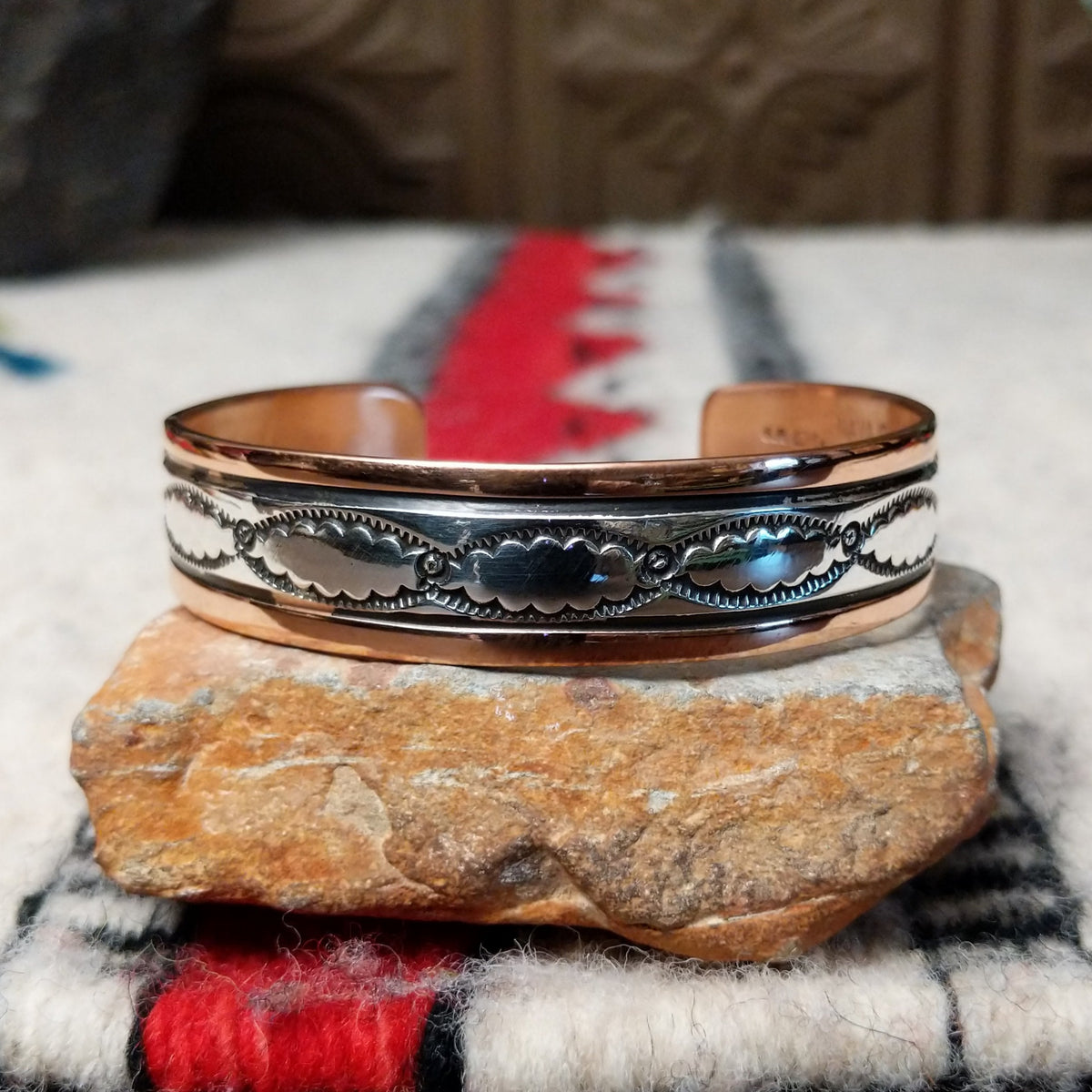 This Copper & Silver Cuff by Robert Little features polished copper borders, a sterling silver center, repeating scalloped hand-stamped motifs, fine textured edges, and dark oxidized accents.