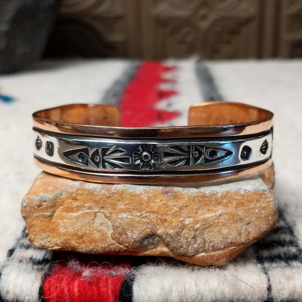 This Copper & Silver Cuff by Robert Little features a polished copper band, sterling silver center, oxidized geometric stamping, and a central floral design.