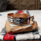 This Copper & Silver Cuff by Robert Little features a polished copper band, sterling silver center, oxidized geometric stamping, and a central floral design; side view.