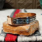 This Copper & Silver Cuff by Robert Little features a polished copper band, sterling silver center, oxidized geometric stamping, and a central floral design; side view.