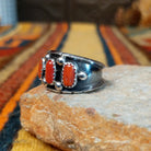 This striking Coral Trio Ring by Reed Charley features three rectangular red-orange coral stones in sawtooth silver bezels, accented by polished silver beads and a wide oxidized silver band; side view.