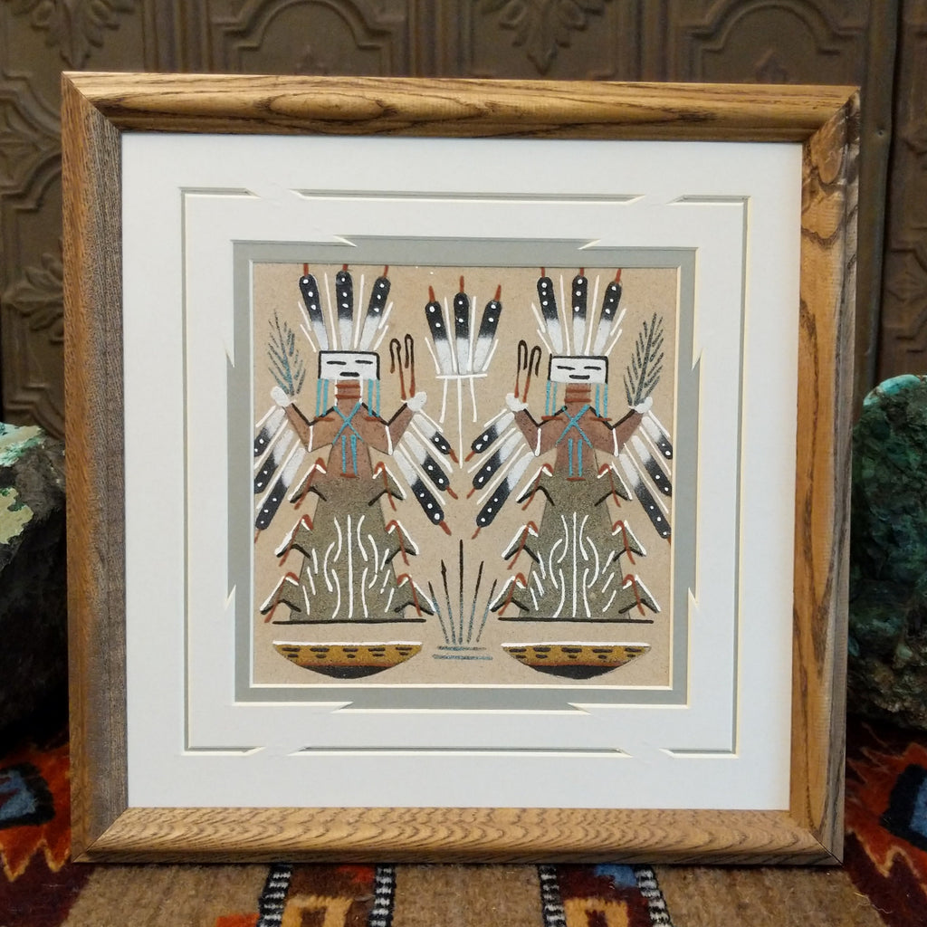 Two Corn People created from sand in multiple colors and set within a wooden frame.