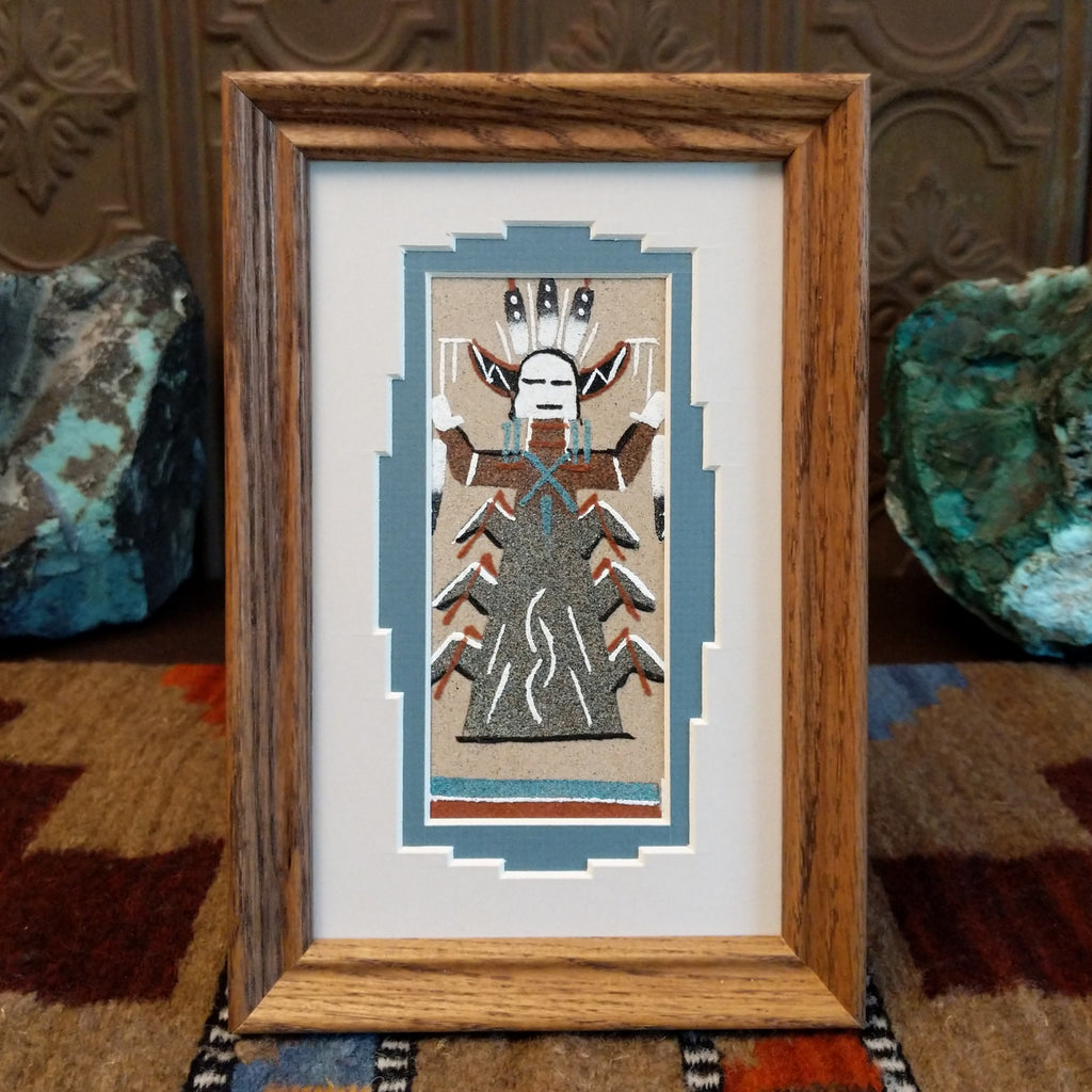 Colorful sand creating a picture of a corn person set within a wooden frame.