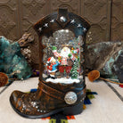 Cowboy Boot Santa Snow Globe featuring cowboy-style Santa with a child, presents, and a snowy evergreen inside a rustic brown Western boot decorated with silver stars and studs.