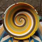 Handwoven earth tone wire art bowl by Bridge for Africa featuring a spiral pattern in recycled wire with golden yellow, olive green, ivory, brown, and tan tones, perfect for home décor or wall display.