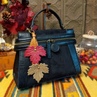 Emma Crossbody Bag by Liz Soto featuring a structured silhouette, mixed-texture panels, top handle, zippered compartment, decorative zipper pulls, and layered leaf-shaped charms.