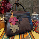 Emma Crossbody Bag by Liz Soto featuring a structured silhouette, mixed-texture panels, top handle, zippered compartment, decorative zipper pulls, and layered leaf-shaped charms.