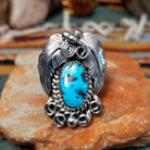 Handcrafted sterling silver ring by Allen Chee featuring an oval natural turquoise cabochon with dark matrix, surrounded by detailed feather motifs, silver scrollwork, twisted wire, and bead accents.