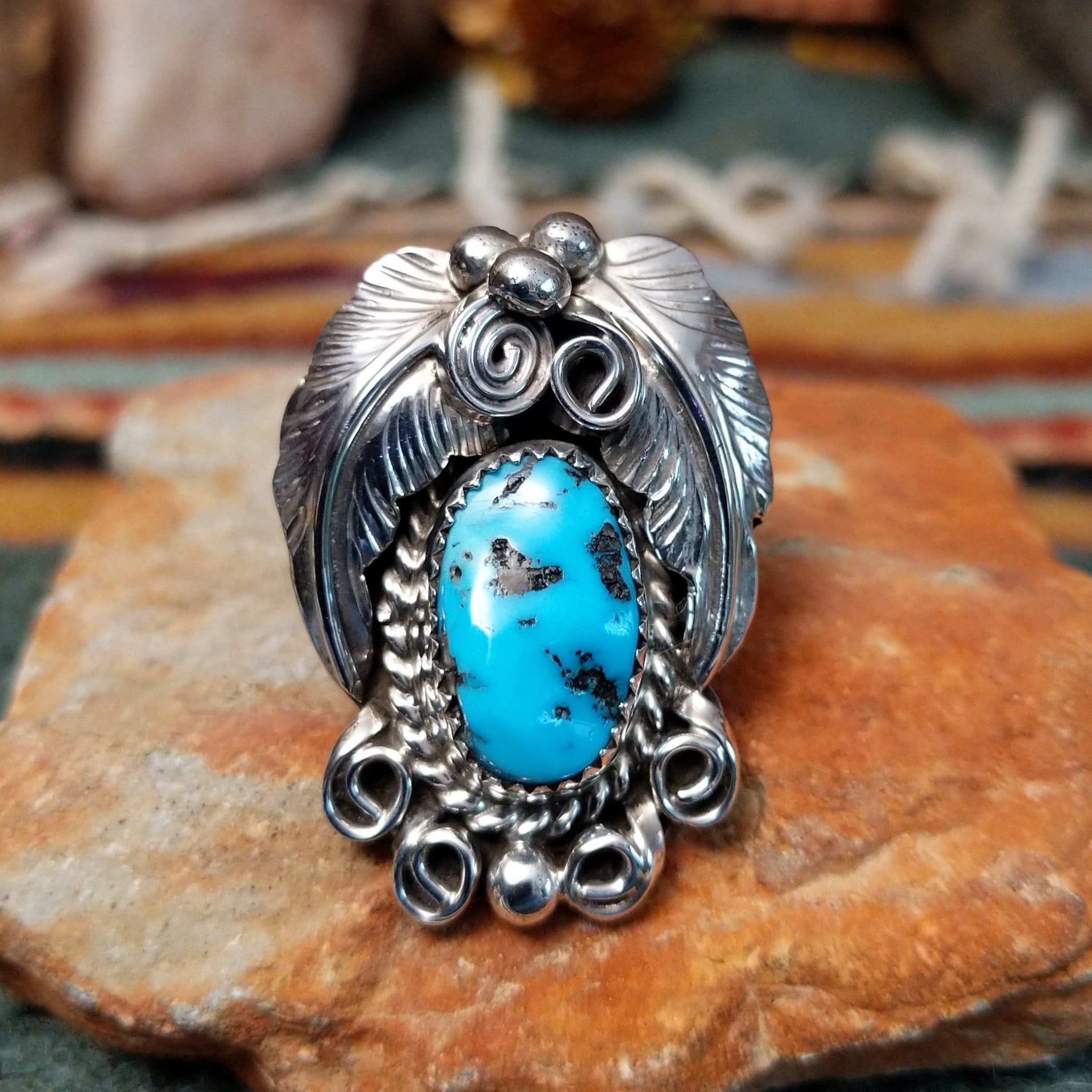 Handcrafted sterling silver ring by Allen Chee featuring an oval natural turquoise cabochon with dark matrix, surrounded by detailed feather motifs, silver scrollwork, twisted wire, and bead accents.