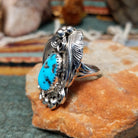 Handcrafted sterling silver ring by Allen Chee featuring an oval natural turquoise cabochon with dark matrix, surrounded by detailed feather motifs, silver scrollwork, twisted wire, and bead accents; side view.