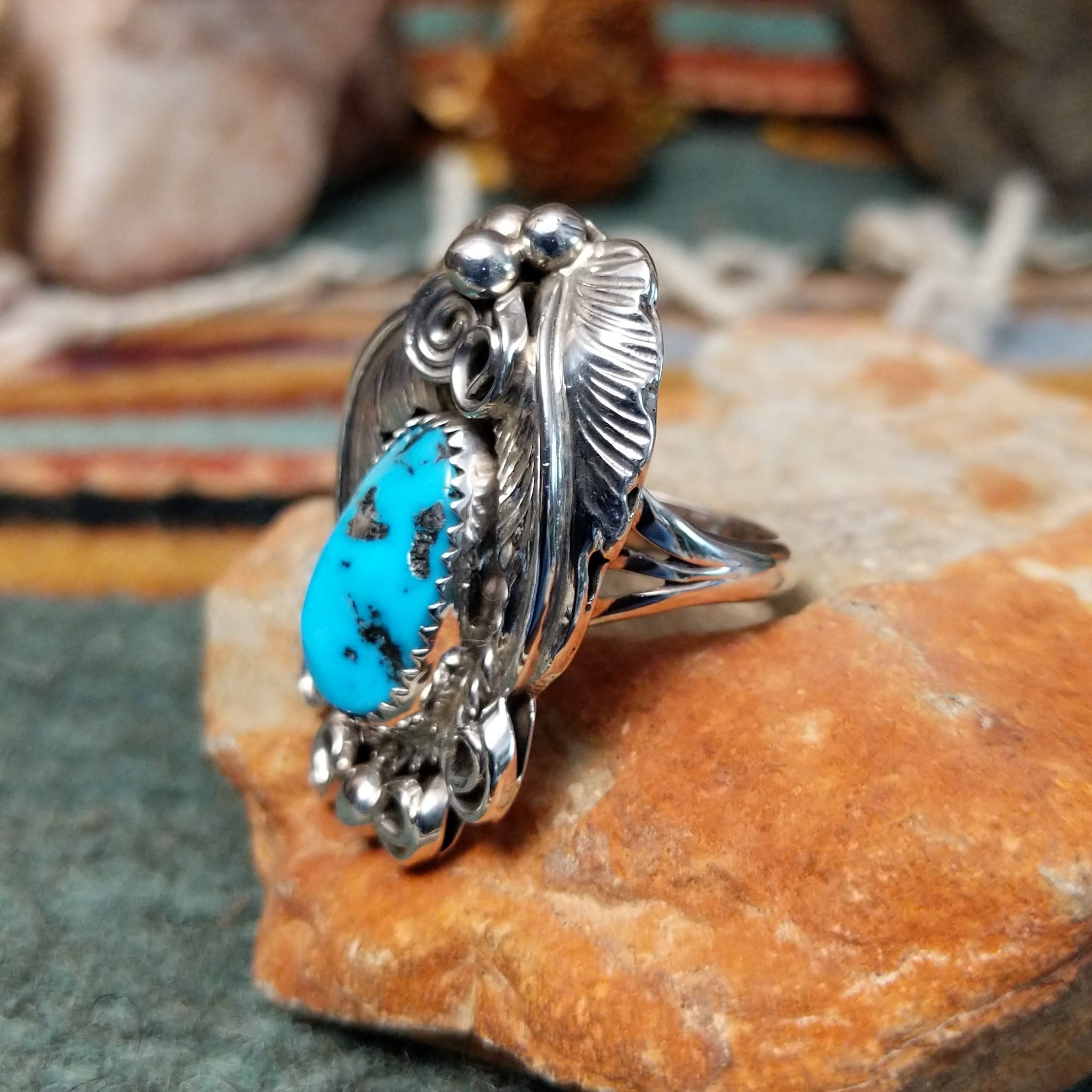 Handcrafted sterling silver ring by Allen Chee featuring an oval natural turquoise cabochon with dark matrix, surrounded by detailed feather motifs, silver scrollwork, twisted wire, and bead accents; side view.