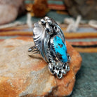 Handcrafted sterling silver ring by Allen Chee featuring an oval natural turquoise cabochon with dark matrix, surrounded by detailed feather motifs, silver scrollwork, twisted wire, and bead accents; side view.