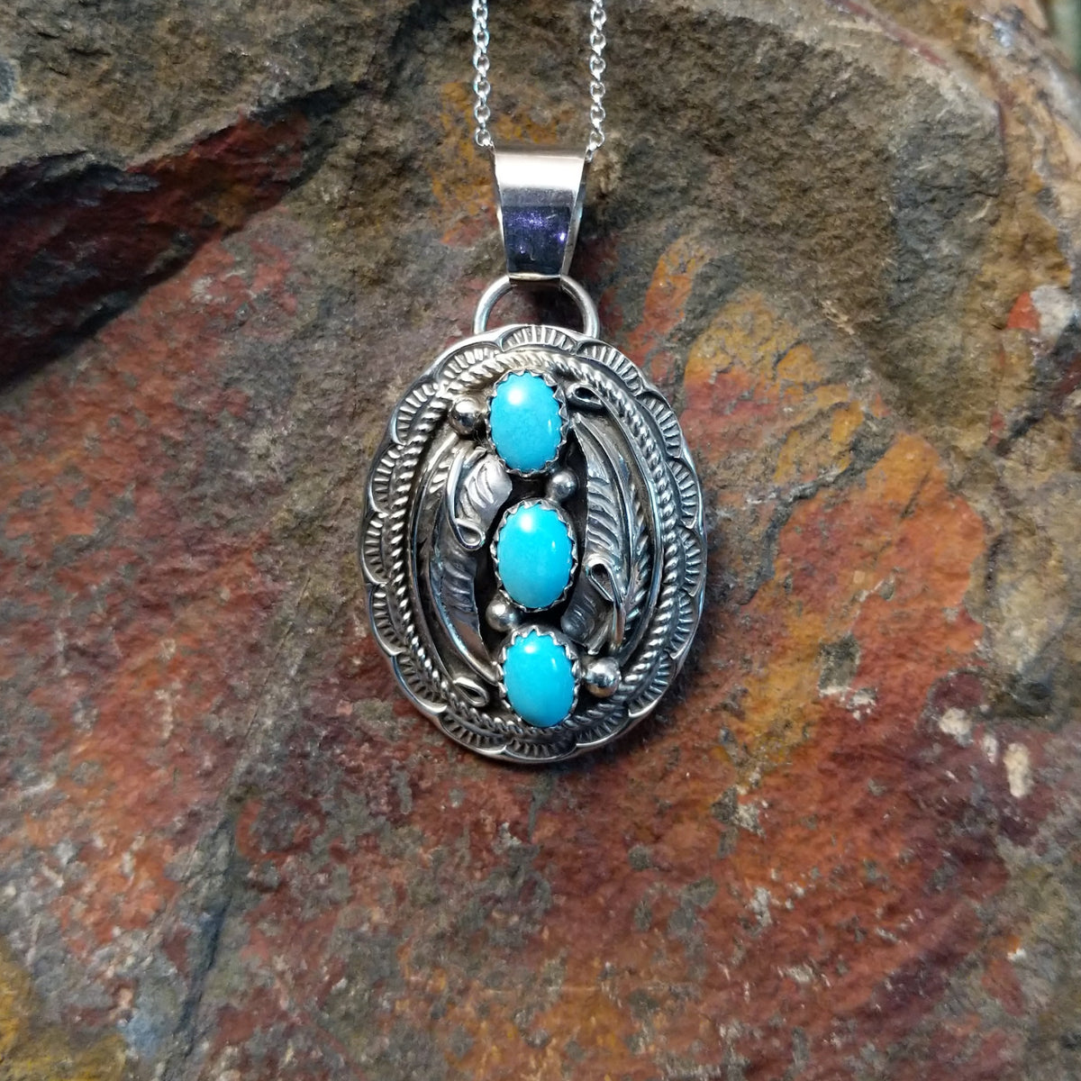 Turquoise pendant by Harrison and Betty Begaye featuring three oval turquoise stones surrounded by detailed sterling silver feathers, twisted rope, stamped borders, and silver accents.