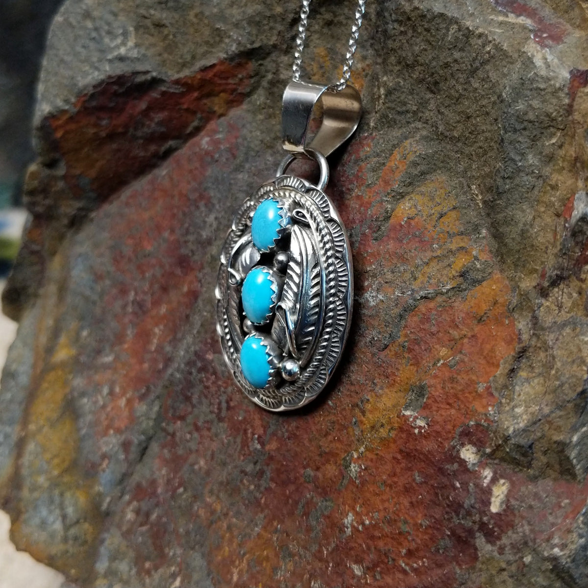 Turquoise pendant by Harrison and Betty Begaye featuring three oval turquoise stones surrounded by detailed sterling silver feathers, twisted rope, stamped borders, and silver accents; side view.