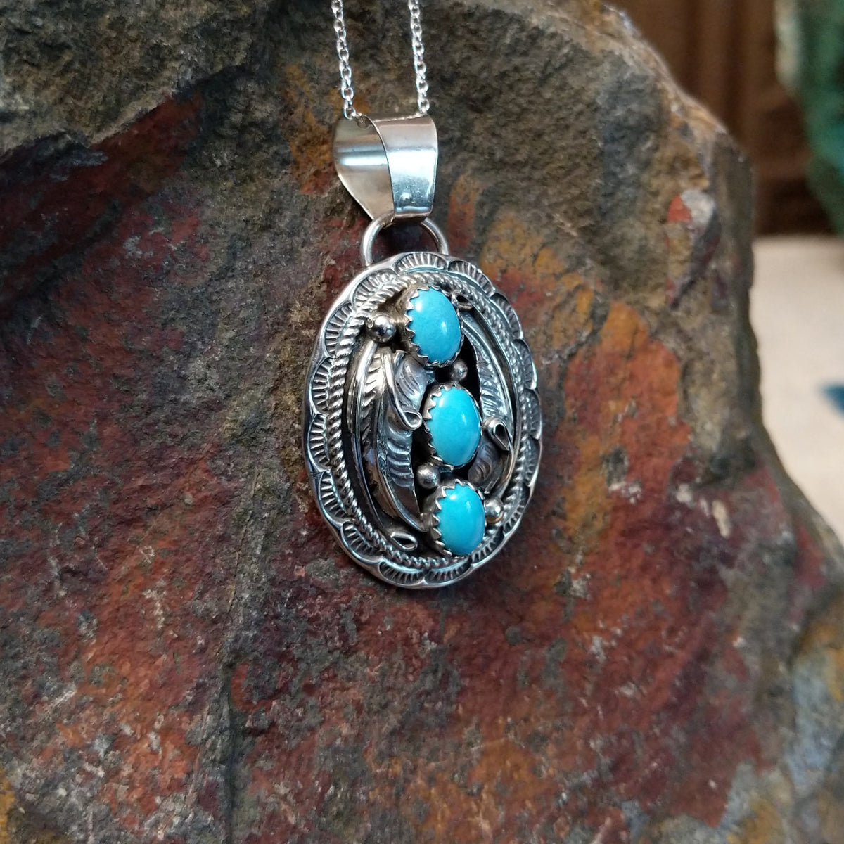Turquoise pendant by Harrison and Betty Begaye featuring three oval turquoise stones surrounded by detailed sterling silver feathers, twisted rope, stamped borders, and silver accents; side view.