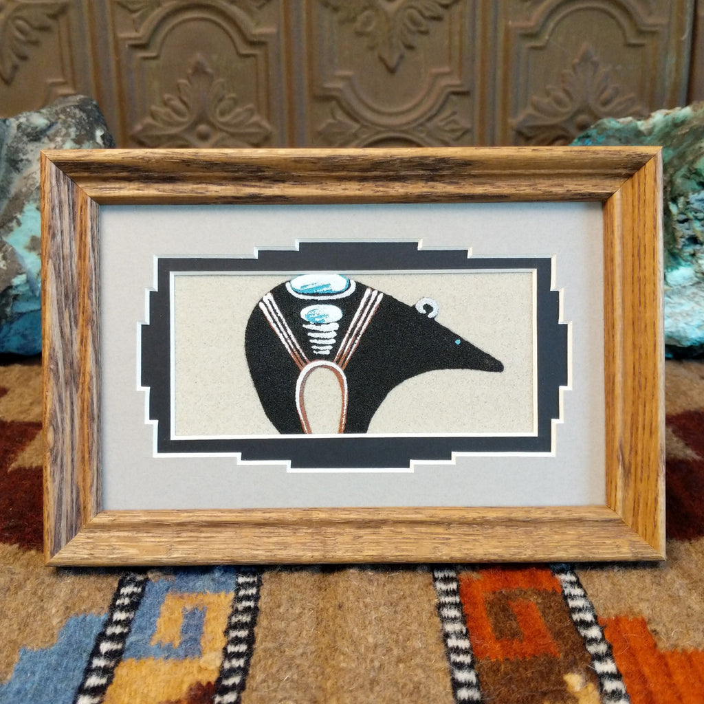 Sand painting of a fetish bear in multiple, contrasting colors set within a wooden frame.