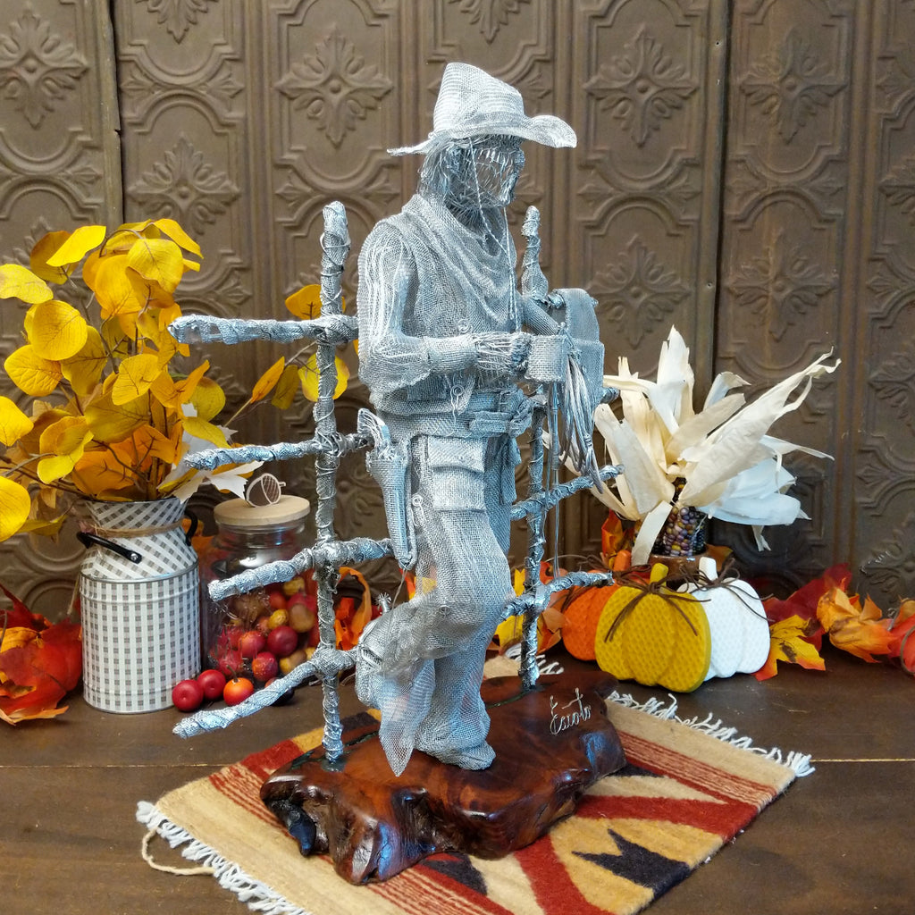 Stainless steel wire and surgical mesh used to create a scene of a cowboy holding a coffee cup and rope, leaning on a fence that is mounted to the mesquite wood base.