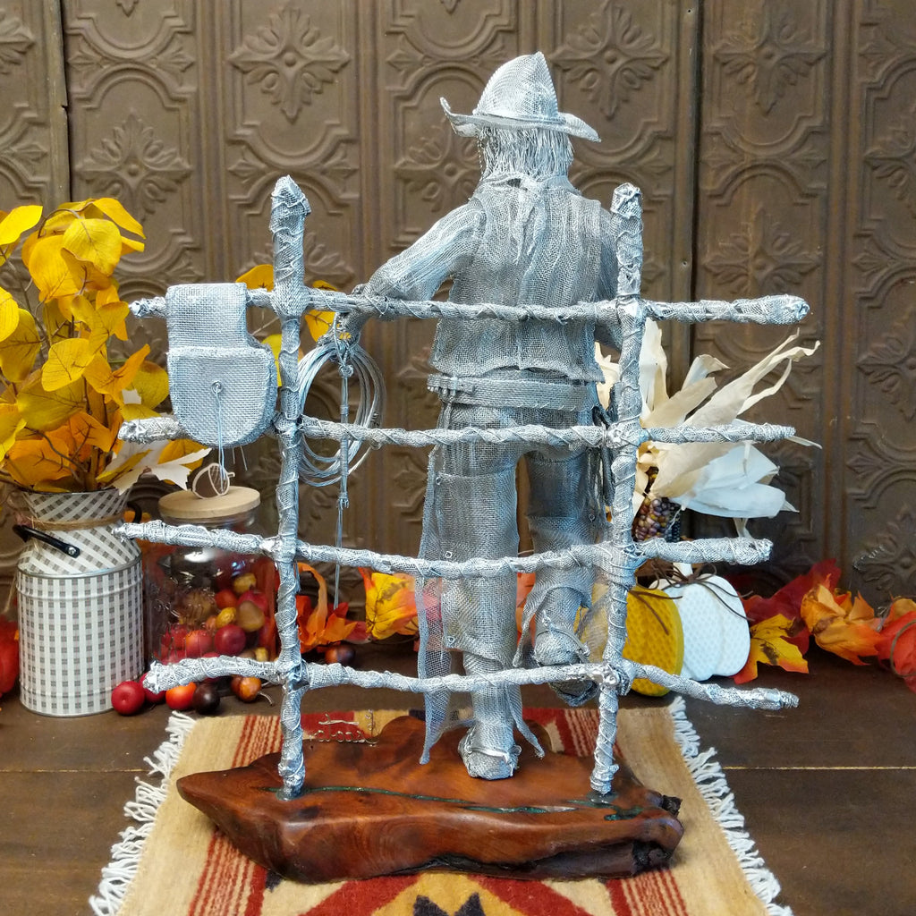 Stainless steel wire and surgical mesh used to create a scene of a cowboy holding a coffee cup and rope, leaning on a fence that is mounted to the mesquite wood base.