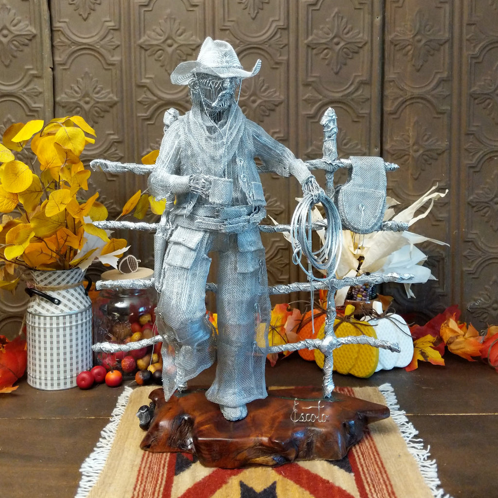 Stainless steel wire and surgical mesh used to create a scene of a cowboy holding a coffee cup and rope, leaning on a fence that is mounted to the mesquite wood base.