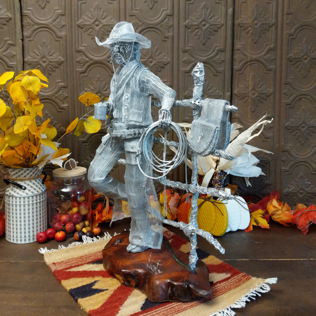 Stainless steel wire and surgical mesh used to create a scene of a cowboy holding a coffee cup and rope, leaning on a fence that is mounted to the mesquite wood base.