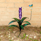 A large purple metal flower with green leaves and a multicolored hummingbird perched above.