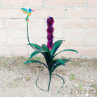 A small purple metal flower with green leaves and a multicolored hummingbird perched above.