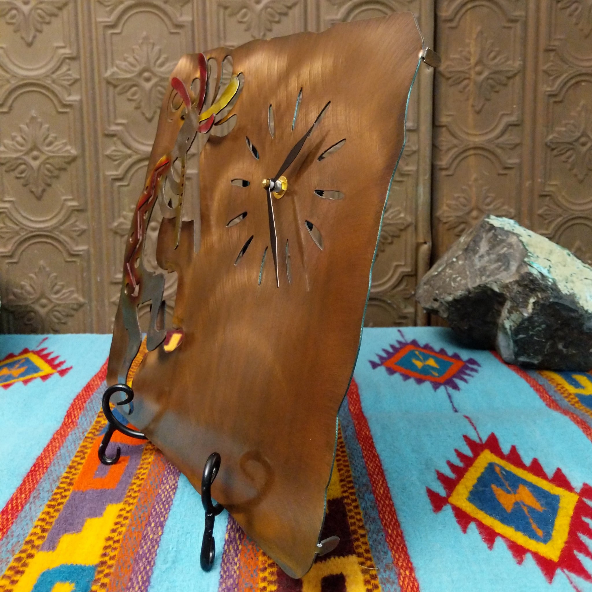 This Crooks Design metal clock features a silver-toned flute-playing Kokopelli with red and yellow Sunset Swirl accents, cutout hour markers, black clock hands, and a warm textured metal background; side view.