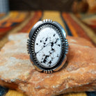 This distinctive White Buffalo Ring by Alfred Martinez features a large oval white stone with black matrix patterns, surrounded by a silver rope border and decorative stamped silver setting.
