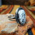 This distinctive White Buffalo Ring by Alfred Martinez features a large oval white stone with black matrix patterns, surrounded by a silver rope border and decorative stamped silver setting; side view.