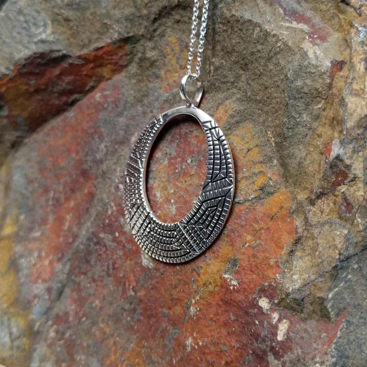 Geometric spiral silver pendant by Elgin Tom featuring an open oval design with intricate stamped patterns, polished silver, and dark oxidized detailing; side view.
