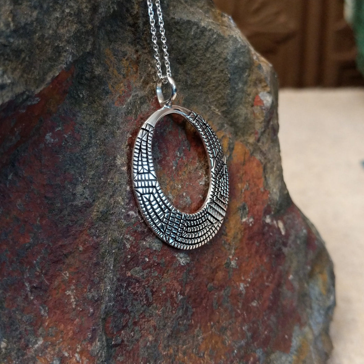 Geometric spiral silver pendant by Elgin Tom featuring an open oval design with intricate stamped patterns, polished silver, and dark oxidized detailing; side view.