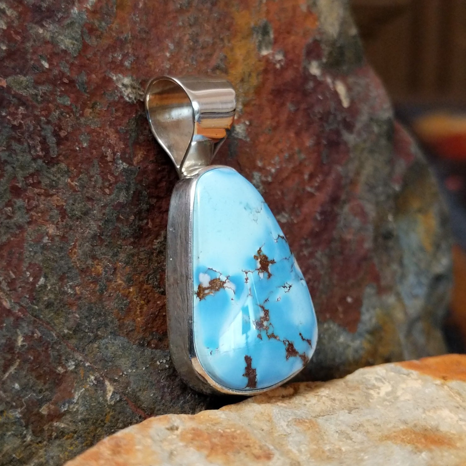 A side view shows the triangular turquoise stone’s brown matrix in a simple silver bezel, with the silver bail gleaming. 