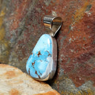 A side view shows the triangular turquoise stone’s brown matrix in a simple silver bezel, with the silver bail gleaming. 
