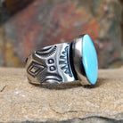 A large, oval Golden Hills turquoise ring by Bernyse Chavez & Lowell Draper, featuring a sky-blue stone with brown matrix veining, set in a wide sterling silver band with a symmetrical sun-ray pattern. The ring rests on a textured sandstone surface with a warm, earthy red background.