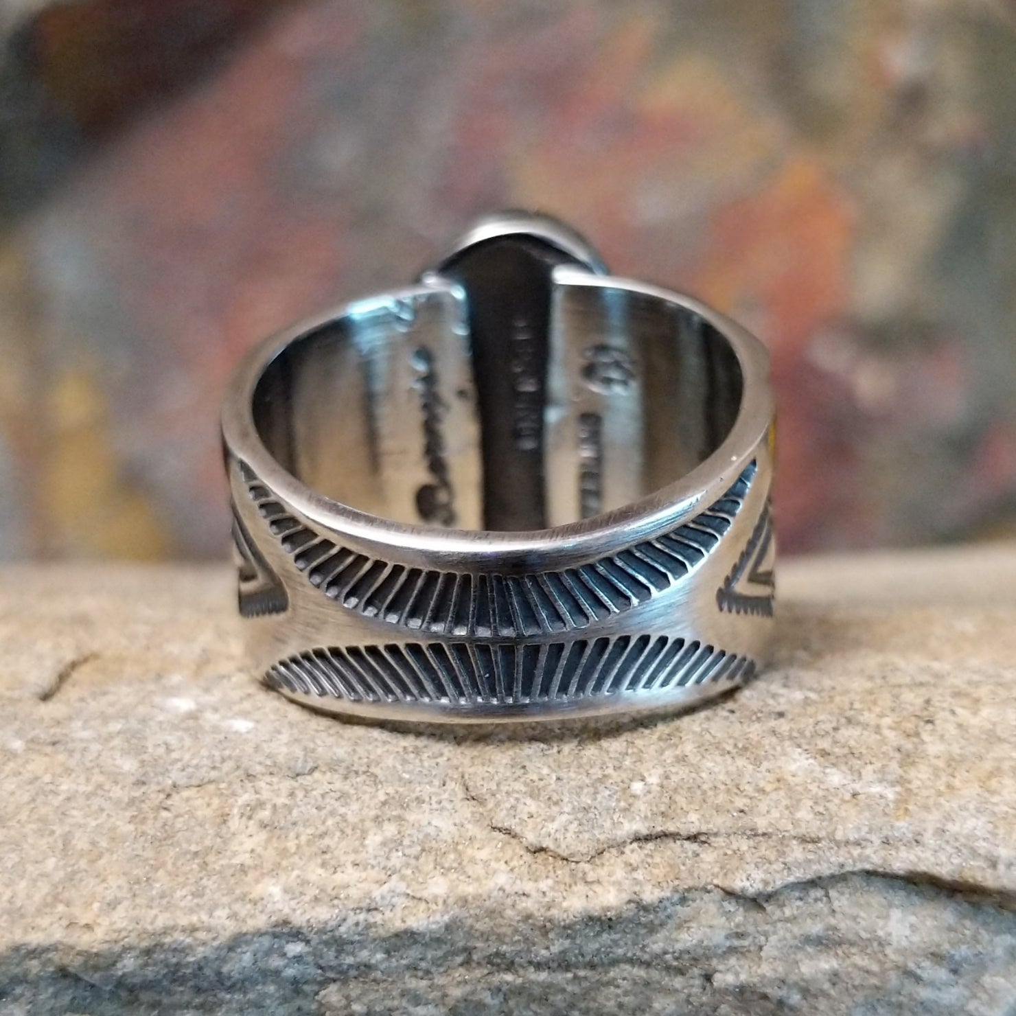 A large, oval Golden Hills turquoise ring by Bernyse Chavez & Lowell Draper, featuring a sky-blue stone with brown matrix veining, set in a wide sterling silver band with a symmetrical sun-ray pattern. The ring rests on a textured sandstone surface with a warm, earthy red background.