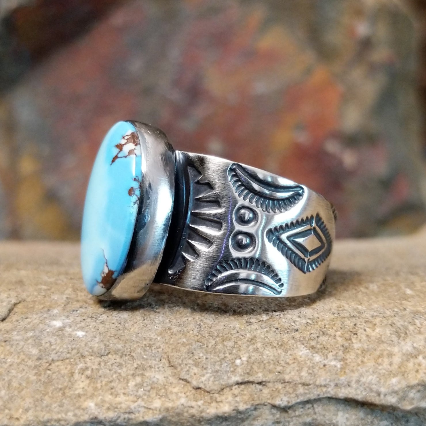 A large, oval Golden Hills turquoise ring by Bernyse Chavez & Lowell Draper, featuring a sky-blue stone with brown matrix veining, set in a wide sterling silver band with a symmetrical sun-ray pattern. The ring rests on a textured sandstone surface with a warm, earthy red background.